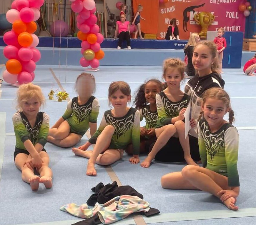 Turnerinnen sitzen mit ihrer Trainerin auf dem Boden und lachen in die Kamera